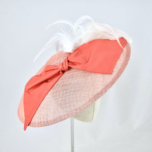 Wide saucer in coral sinamay with coral silk bow and white feathers on a bandeau base. 3/4 front view.