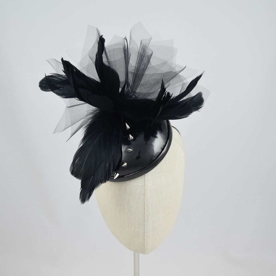 Cocktail hat in wet look vinyl with feathers, tulle, and studs. Front view.