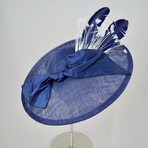 Royal blue sinamay saucer with silk bow and hand dyed feathers on a bandeau base. Top front view.