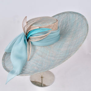 Turquoise and silver sinamay hat with wide, sweeping brim trimmed with turquoise silk chiffon bow and sinamay leaves.  Front view.